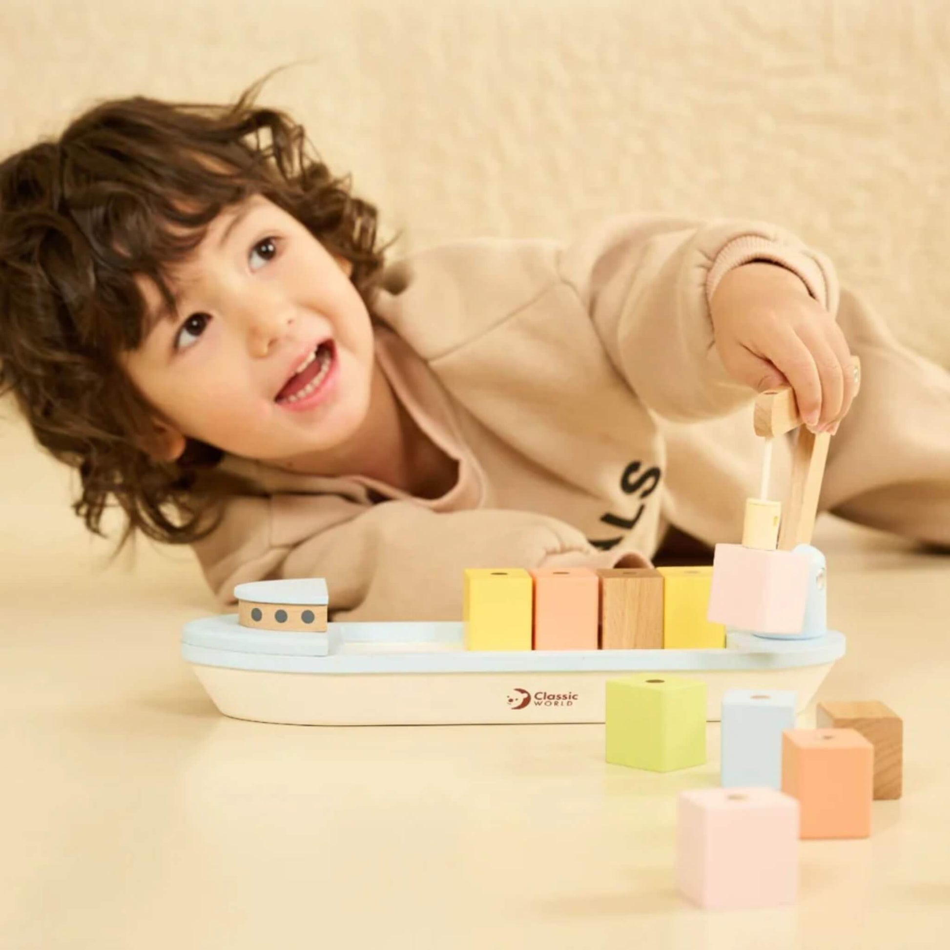 Smiling child lifting a pink wooden block with the crane arm of a cargo ship toy.