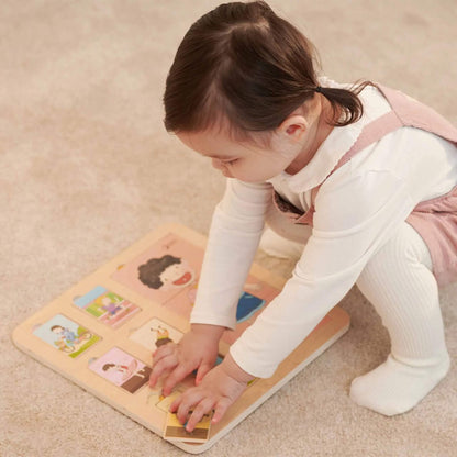 Toddler in white and pink clothing crouched over a wooden puzzle, carefully arranging pieces on the board.