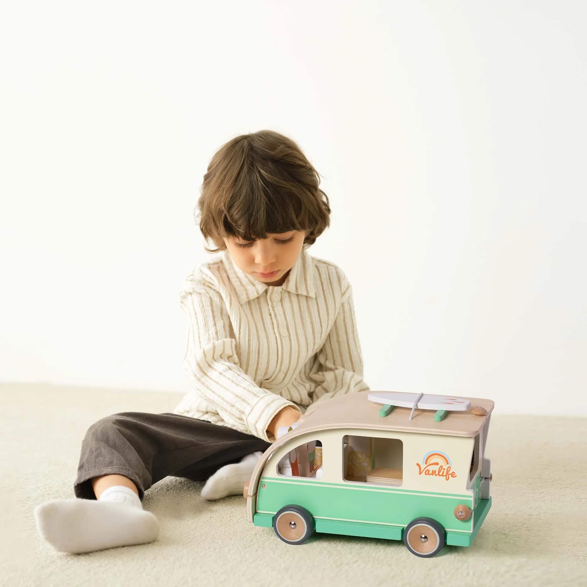 A young child sitting on the floor playing with a wooden toy campervan, interacting with its removable parts.