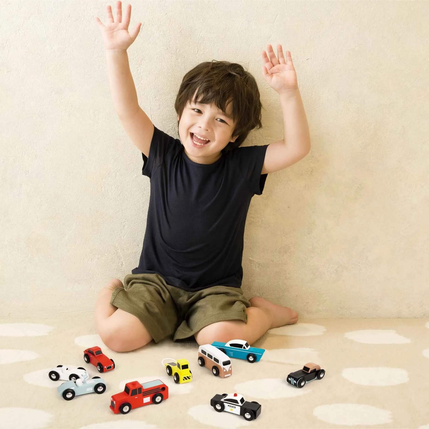 Smiling child sitting on the floor with hands raised, surrounded by an assortment of wooden toy cars, including a police car, fire truck, camper van, and sports cars.