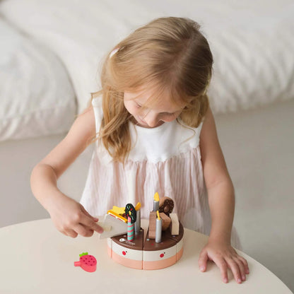 A child playing at a table with the wooden toy chocolate cake, using the wooden knife to serve pieces, surrounded by colourful wooden toppings.