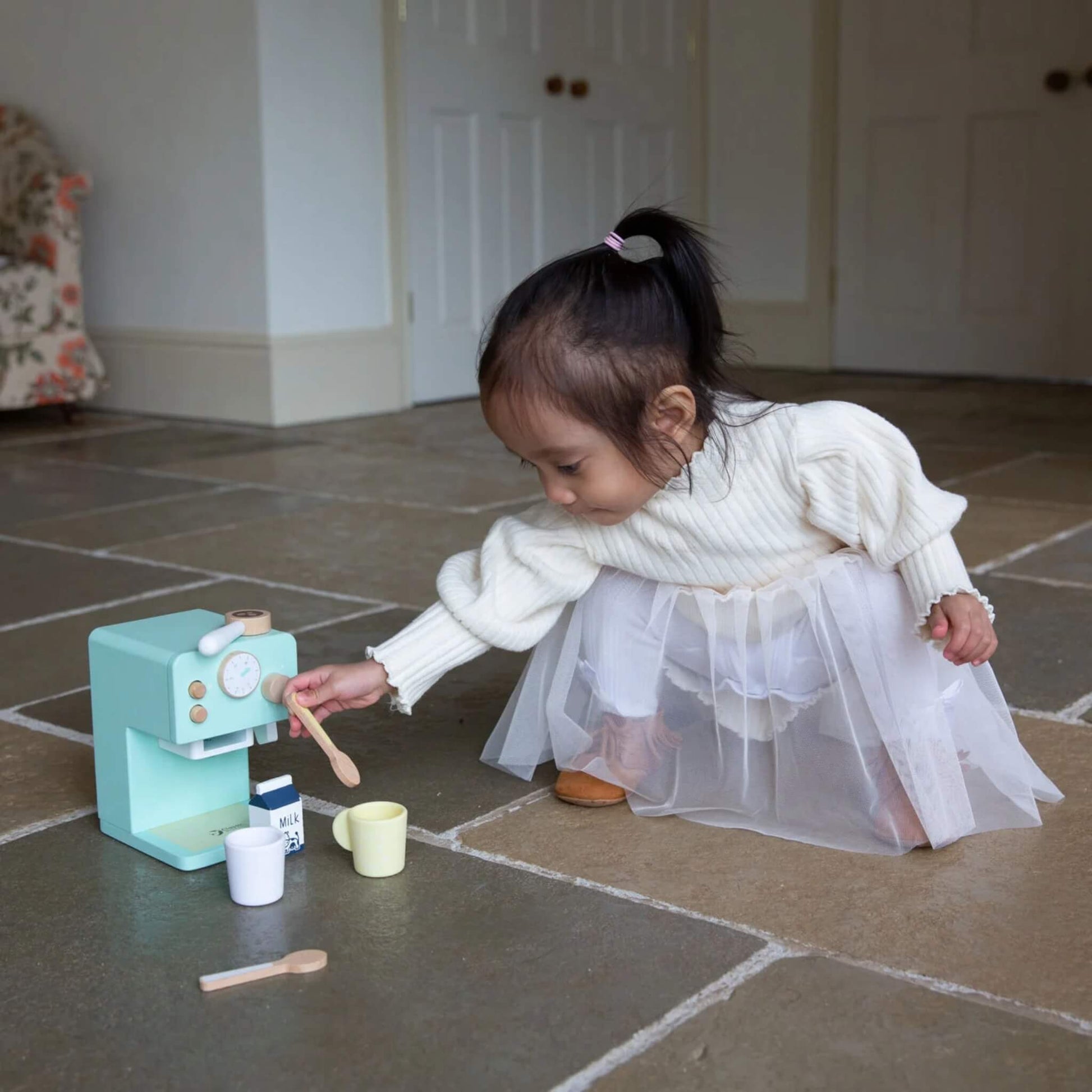 Toddler in a white dress crouching on a stone floor, reaching out to play with the wooden coffee maker set and accessories.