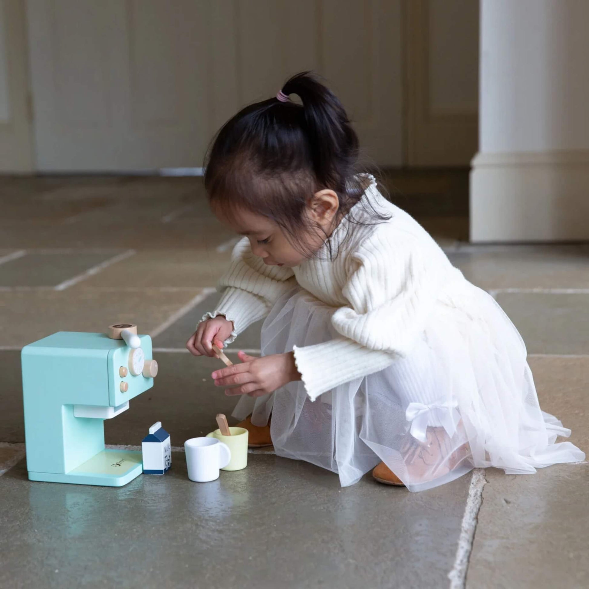 Toddler stirring one of the mugs while sitting in front of the wooden coffee machine, fully engaged in pretend play.