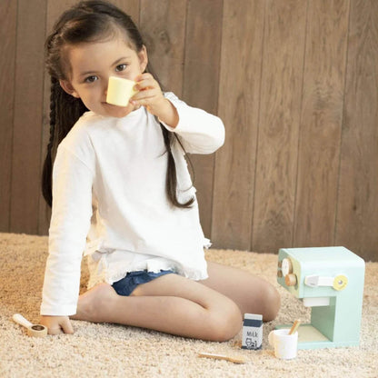 Young child sitting on a soft carpet, lifting a yellow mug in a playful toast next to the mint green wooden coffee machine.