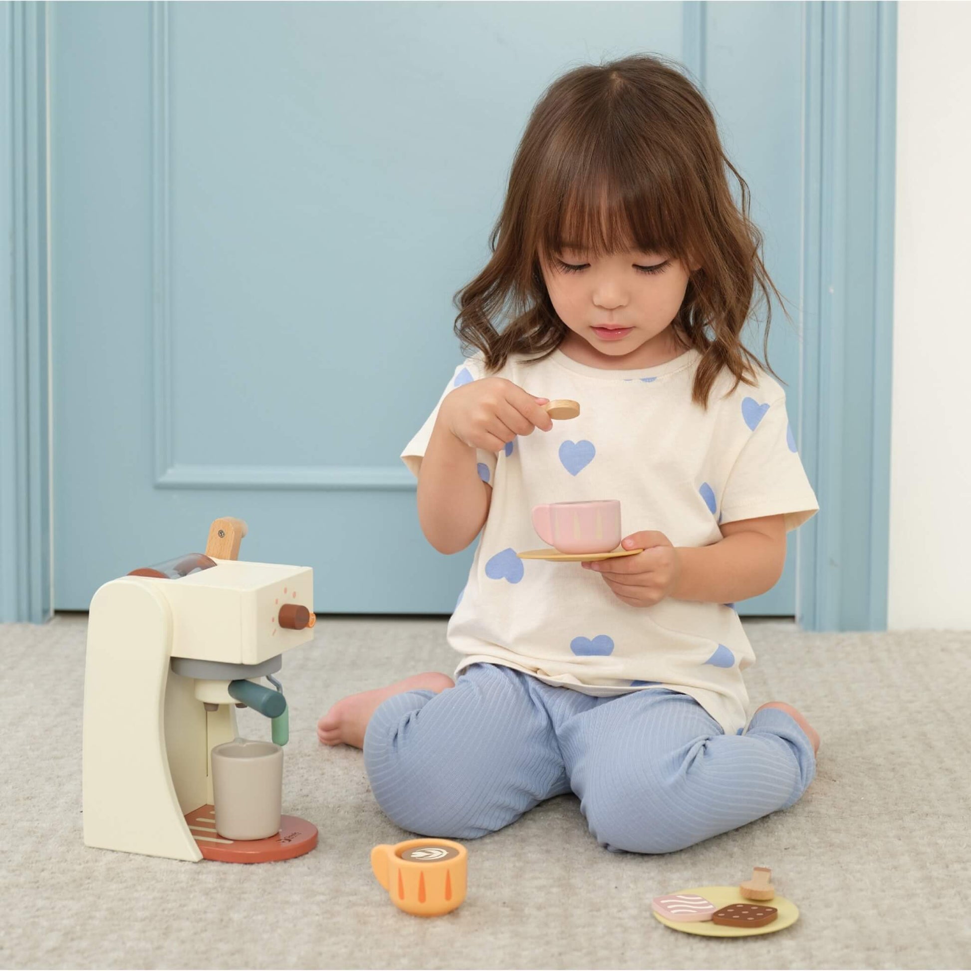A young child sitting on a carpet using a wooden toy coffee machine, holding a spoon and cup while preparing pretend drinks.