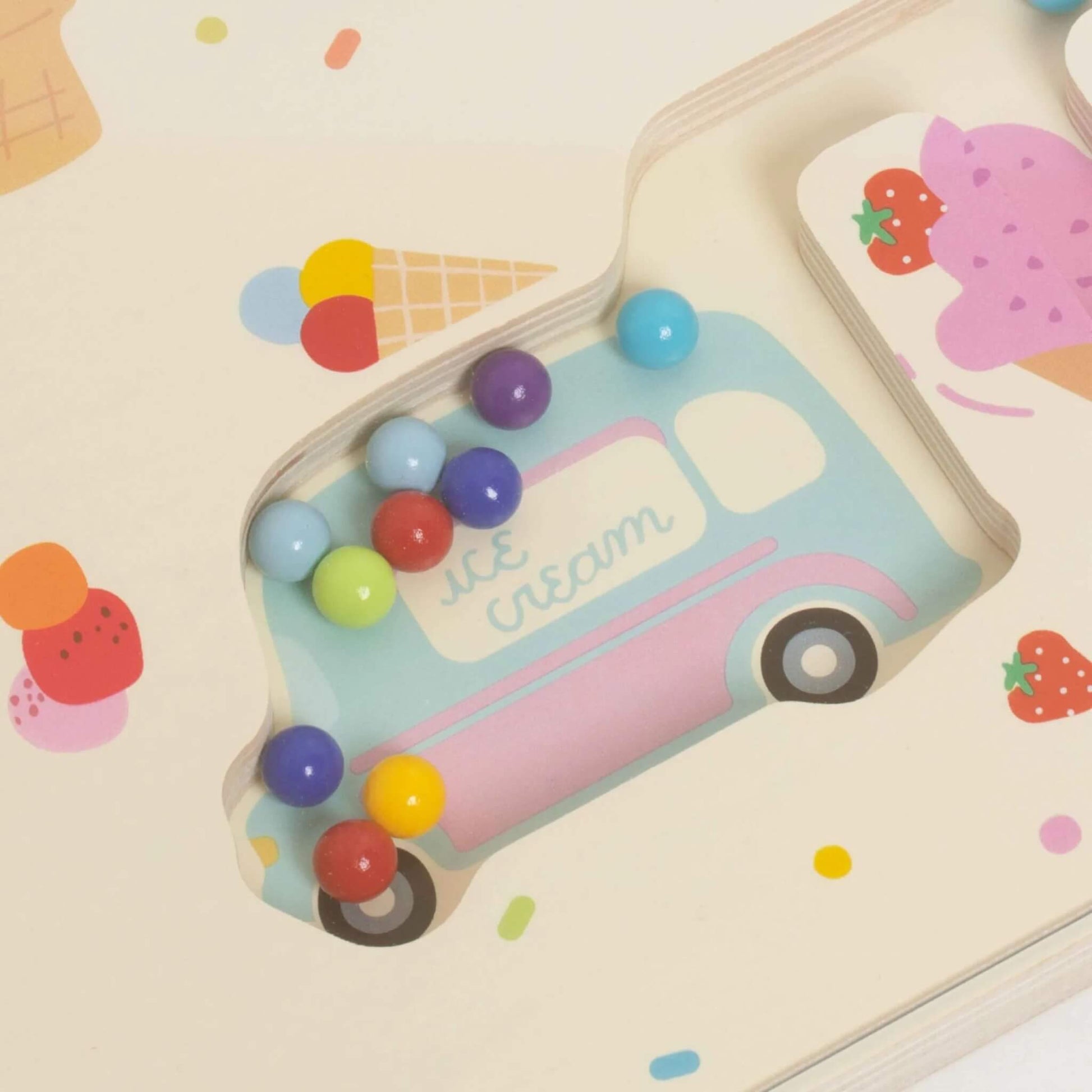 Detail of a wooden magnetic maze with multicoloured balls collected inside a pastel blue ice cream van shape on the board.