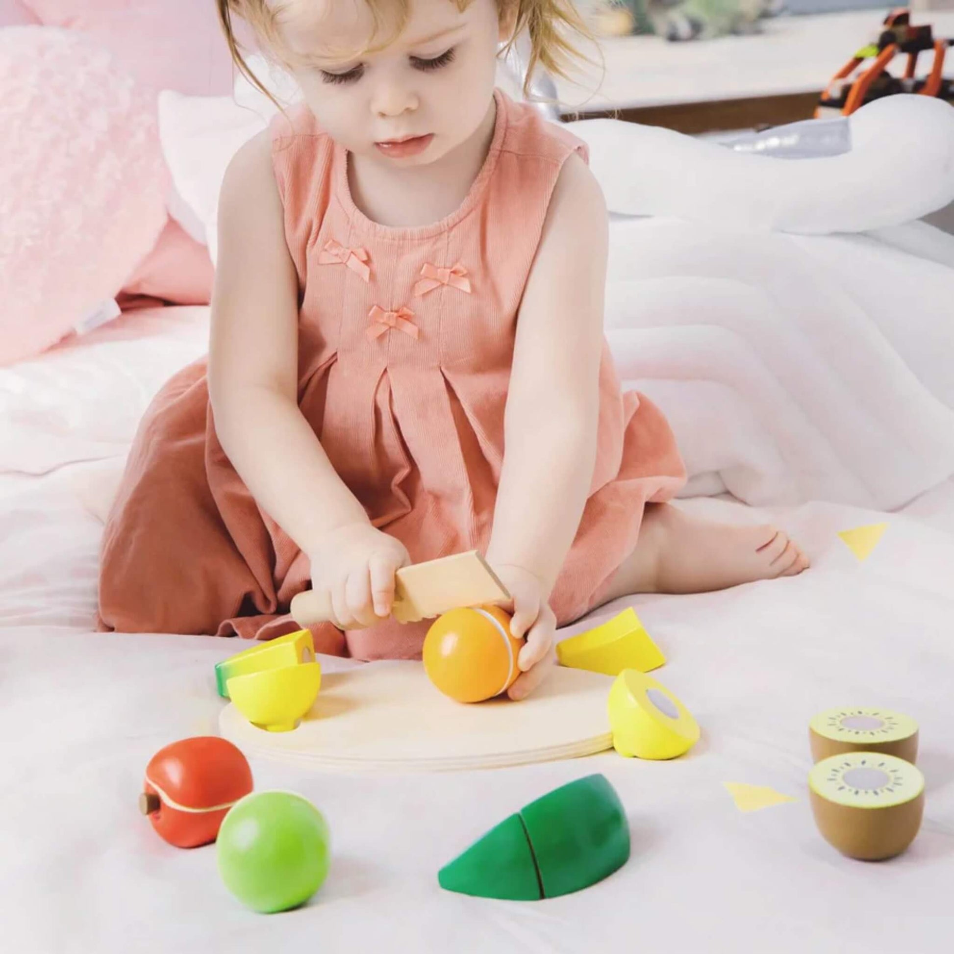 Toddler in a peach-coloured dress sitting on a soft bed, using a wooden toy knife to cut a wooden orange fruit on a small cutting board surrounded by colourful wooden fruits.