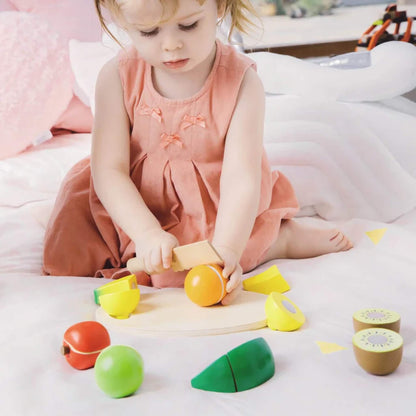 Toddler in a peach-coloured dress sitting on a soft bed, using a wooden toy knife to cut a wooden orange fruit on a small cutting board surrounded by colourful wooden fruits.