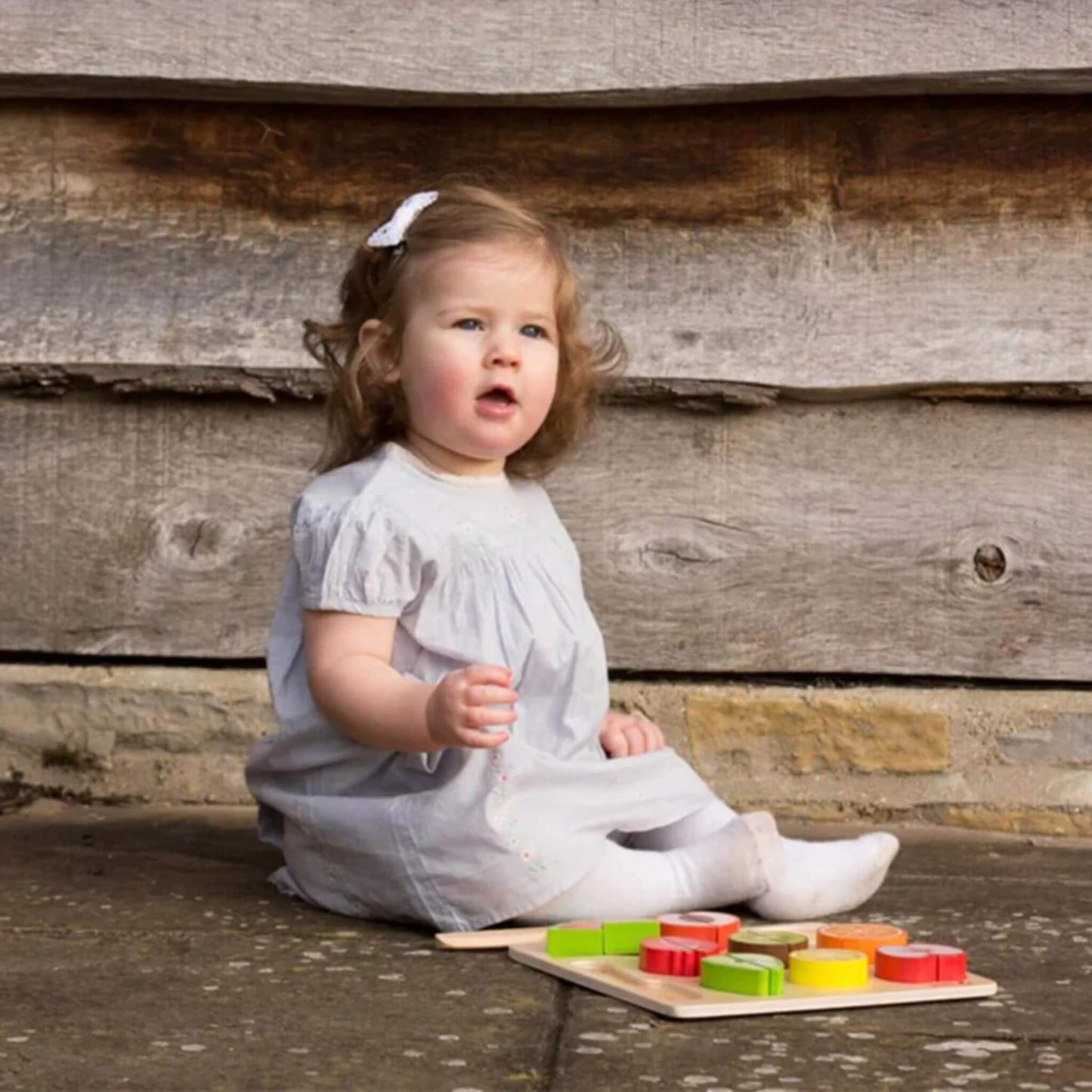 Toddler in a pale grey dress with a white hair clip sitting on outdoor stone flooring, next to a wooden toy cutting set with colourful fruit pieces neatly arranged on a wooden cutting board.