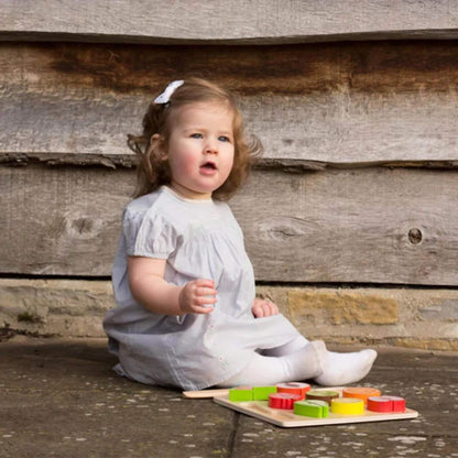 Toddler in a pale grey dress with a white hair clip sitting on outdoor stone flooring, next to a wooden toy cutting set with colourful fruit pieces neatly arranged on a wooden cutting board.