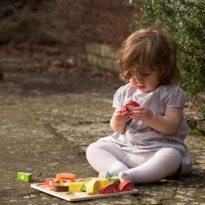 Toddler in a pale grey dress sitting cross-legged on outdoor stone flooring, closely examining two red wooden apple halves from a toy cutting set with scattered colourful fruit pieces nearby.