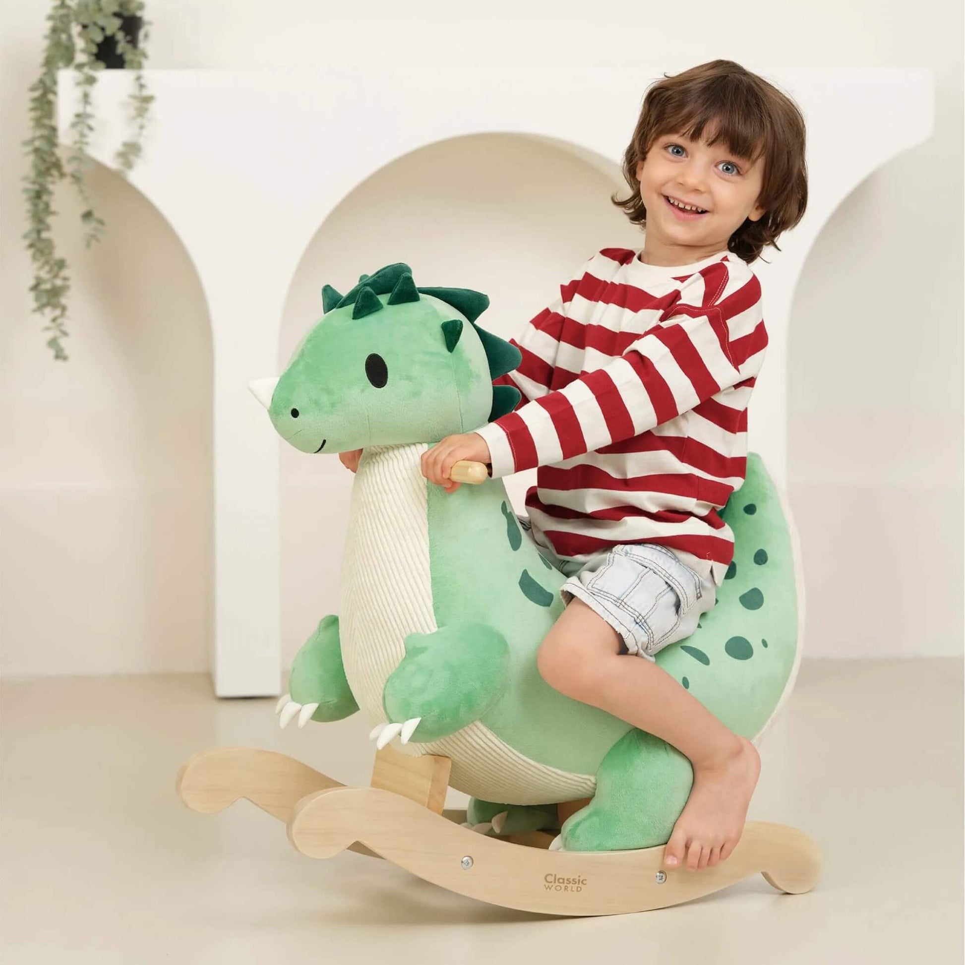 A young child smiling and riding on a plush green dinosaur rocker with wooden handles and a wooden base, indoors on a light floor.