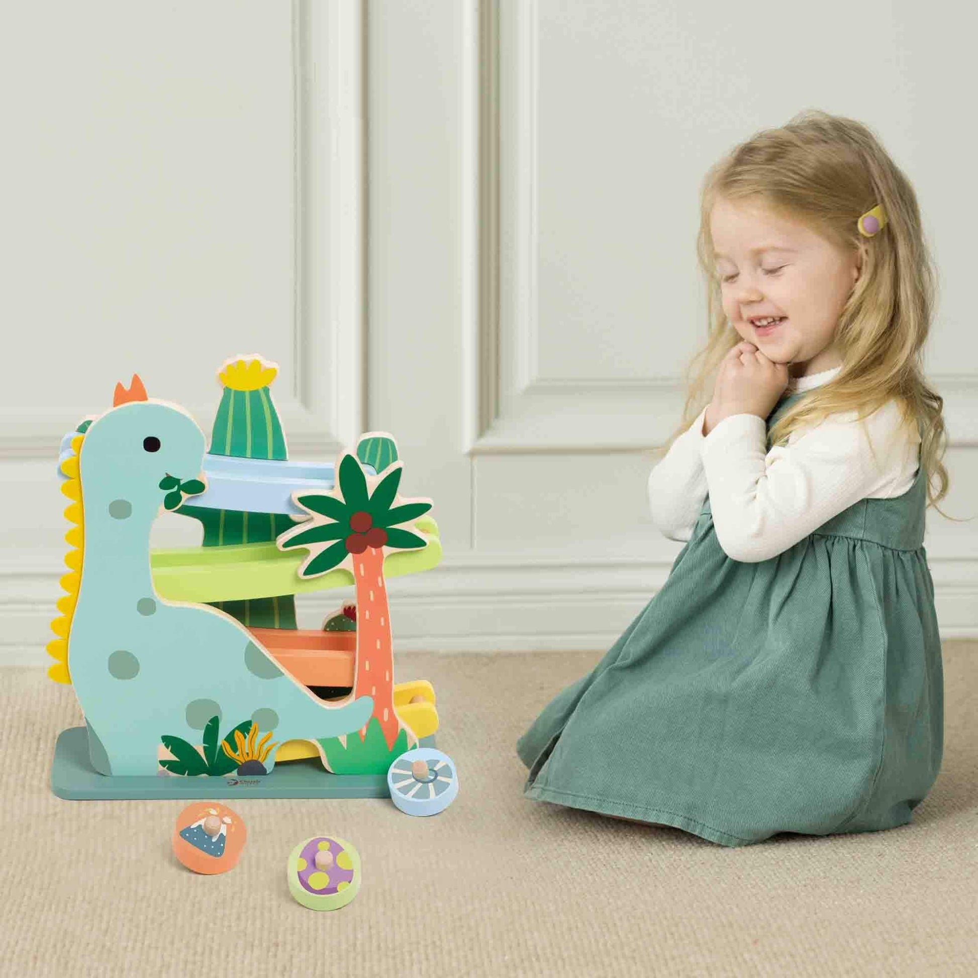 Young girl in a green dress sitting on a carpet, smiling at a wooden dinosaur ramp toy with colourful tracks and three wooden rolling discs in front.