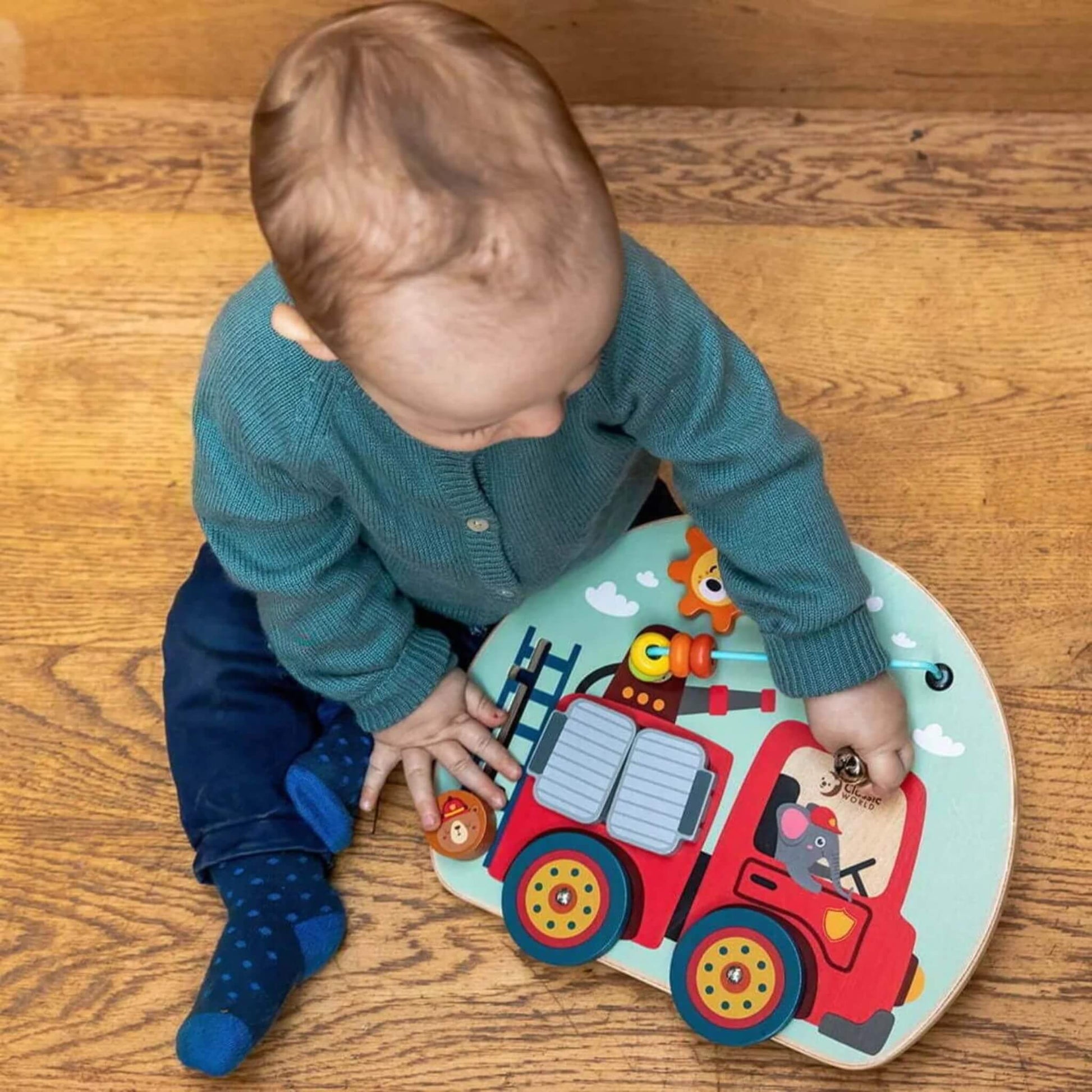 Baby sitting on wooden floor, using fingers to explore the busy board’s ladder and bell.