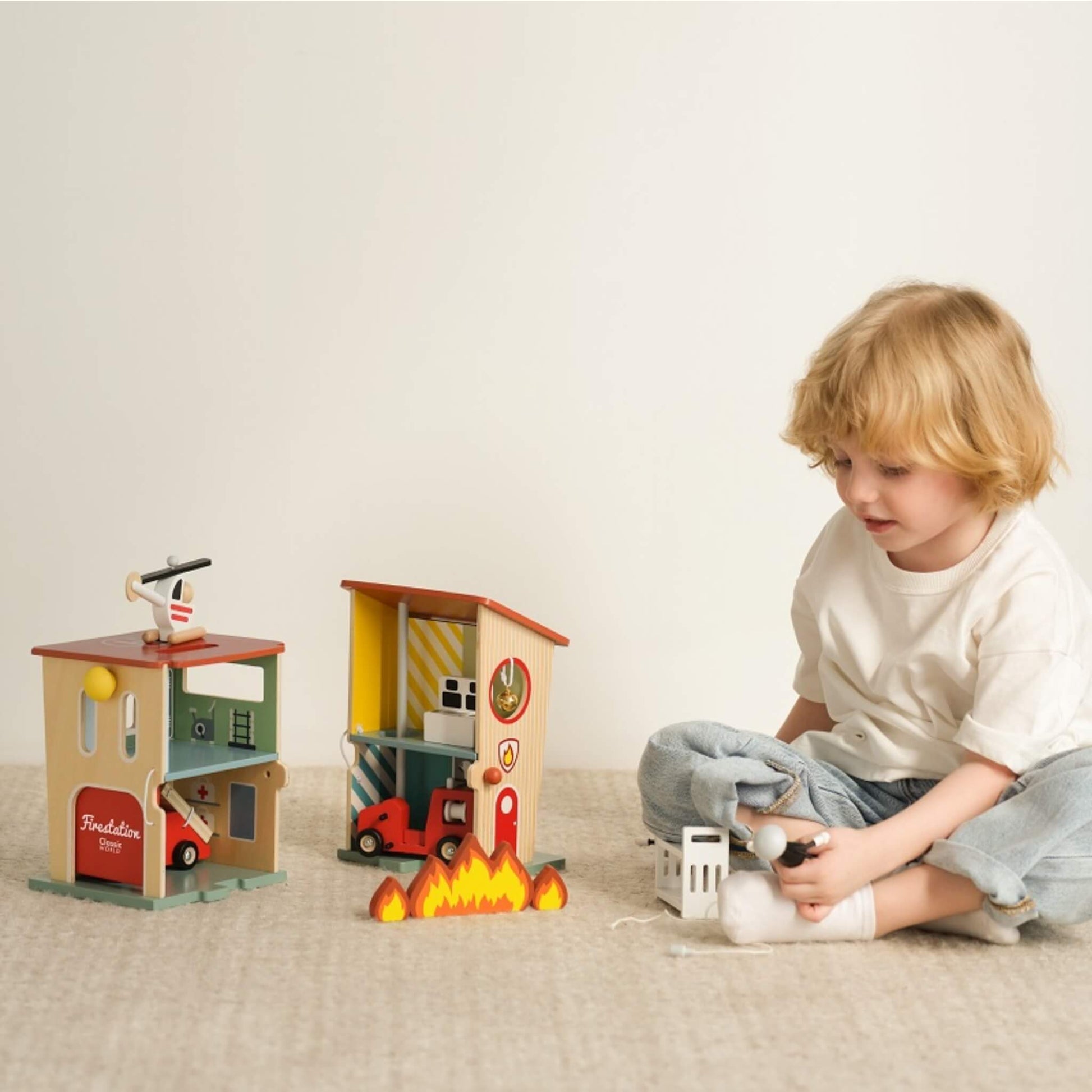 A young child sitting on a carpet playing with a wooden fire station, holding a firefighter figure beside vehicles and flames.