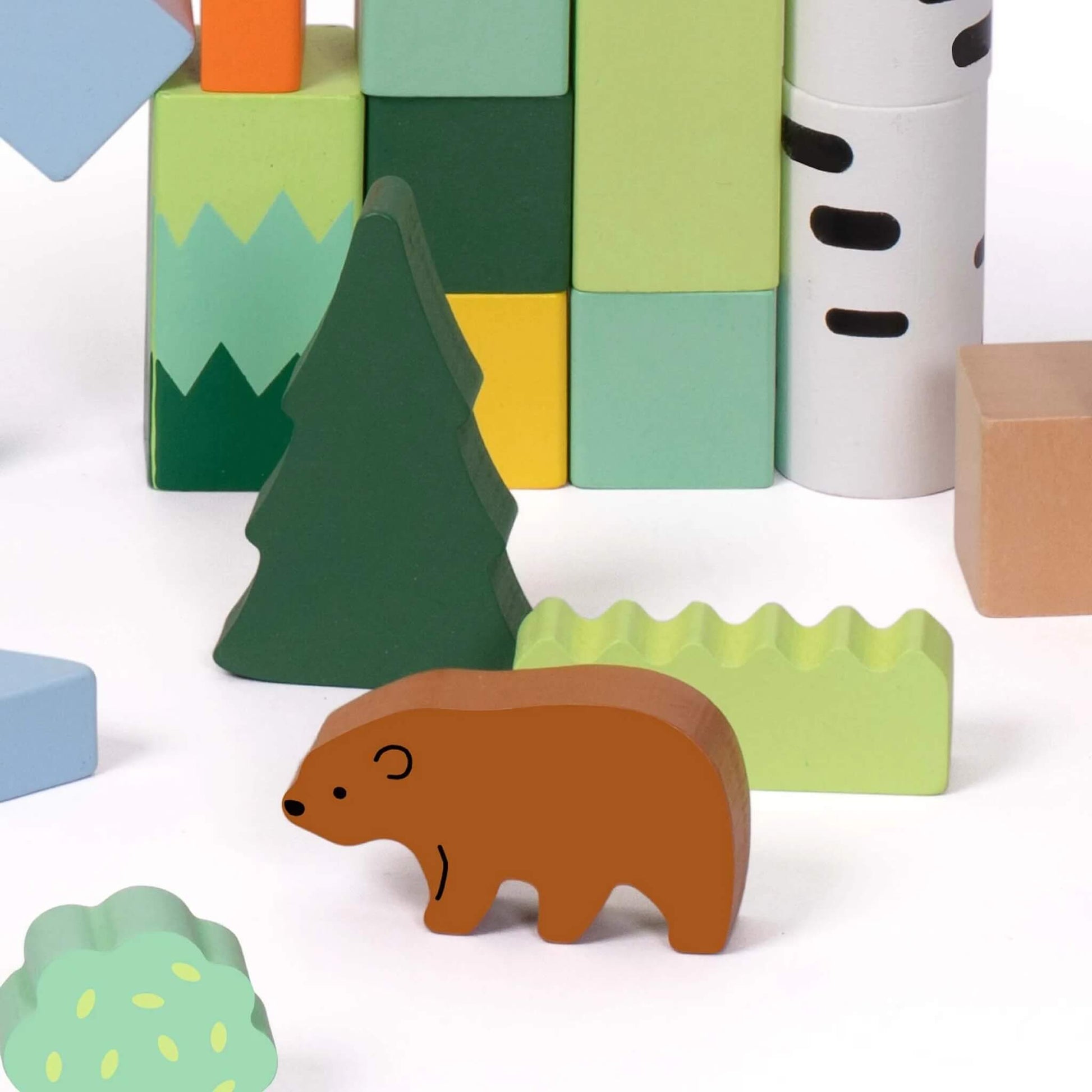 Close-up of a brown bear wooden figure standing among forest-themed blocks, including a pine tree, light green bush, and a stack of pastel building blocks.