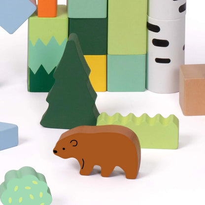 Close-up of a brown bear wooden figure standing among forest-themed blocks, including a pine tree, light green bush, and a stack of pastel building blocks.