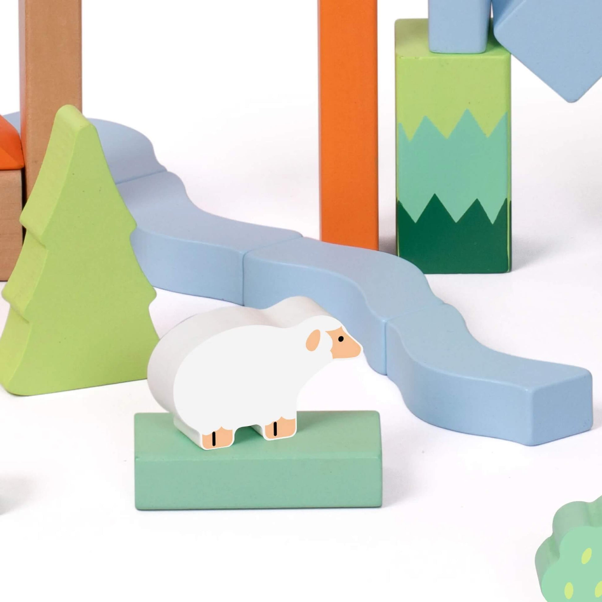 Close-up of a white wooden sheep placed on a light green block, positioned beside light blue wavy blocks resembling a river and surrounded by soft green trees.