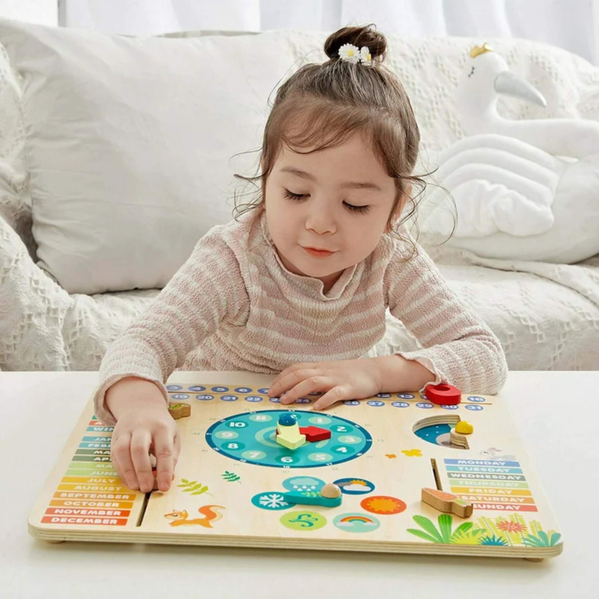 Child using a wooden calendar board with movable weather pieces, adjustable clock hands and colourful month and weekday sections on a white table.