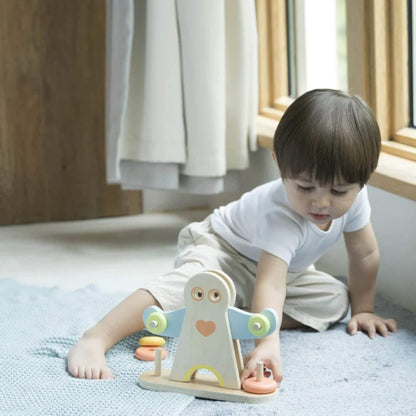 A young child sitting on a soft blue textured blanket indoors, wearing a white shirt and beige pants, reaching out to place a wooden ring on the Hercules toy’s arm.