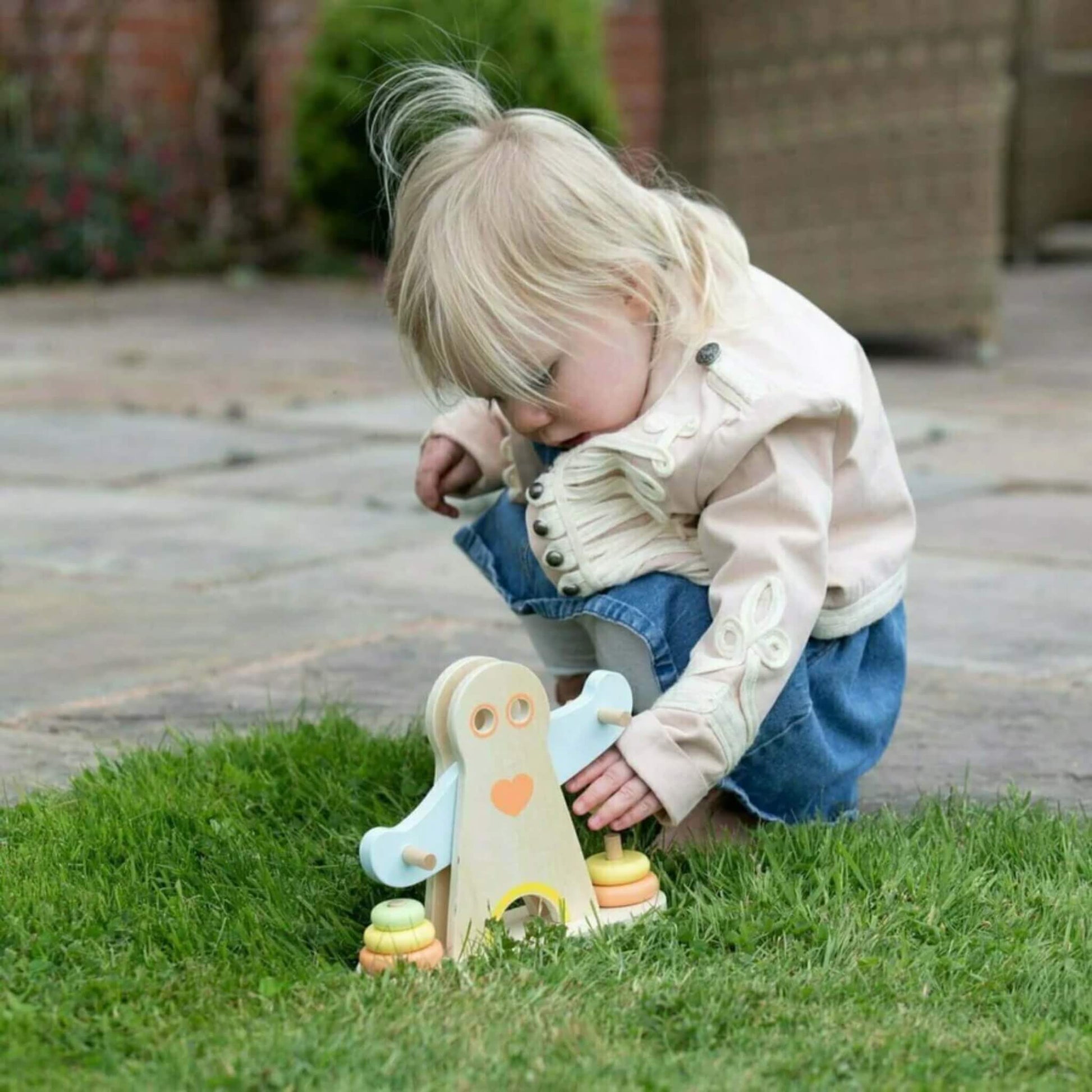 Small child outdoors on green grass, wearing a cream jacket and denim skirt, crouching and using both hands to adjust the pastel-coloured stacking rings on a wooden toy figure shaped like a strong man with outstretched blue arms.