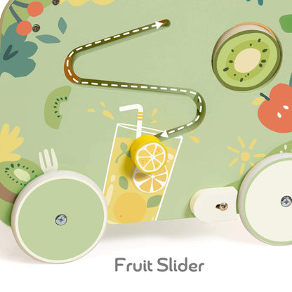 Close-up of a green wooden panel with a winding maze track, showing a yellow wooden lemon slice sliding along a printed fruit drink design, next to a kiwi disc and red apple illustration.