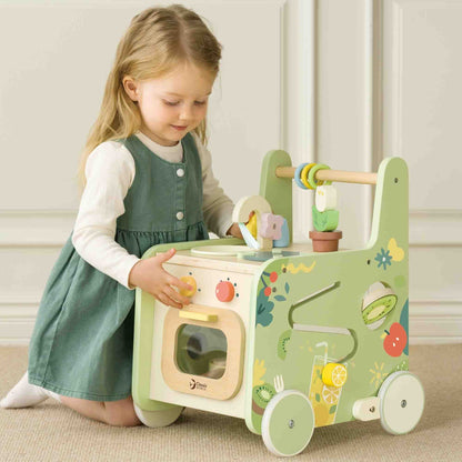 Young child in a green dress turning the oven knob on a pastel wooden play kitchen walker, focused on pretend play.