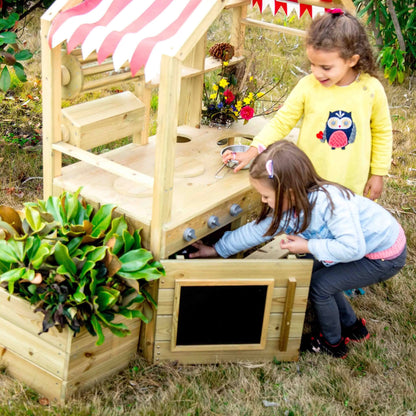 Rear view of two young girls engaging with a wooden outdoor play kitchen, under a striped canopy with garden plants nearby.