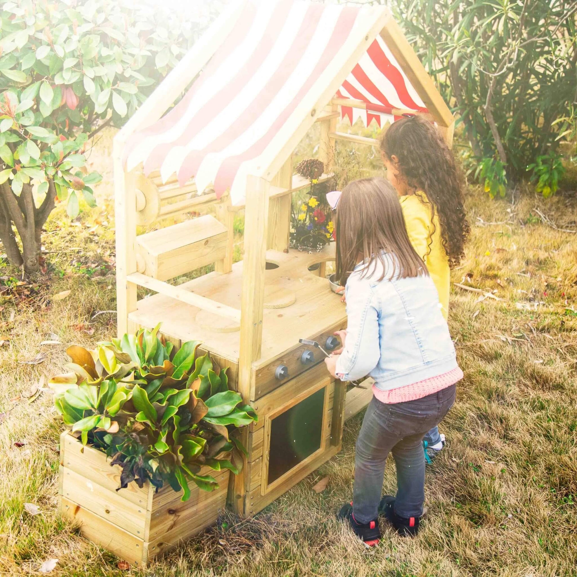 Two children playing at a wooden outdoor play kitchen, turning knobs and exploring the oven, surrounded by plants and flowers in the garden.