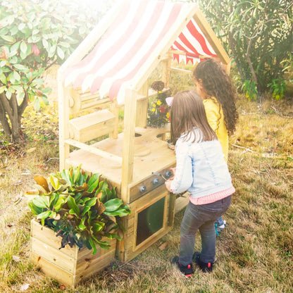 Two children playing at a wooden outdoor play kitchen, turning knobs and exploring the oven, surrounded by plants and flowers in the garden.