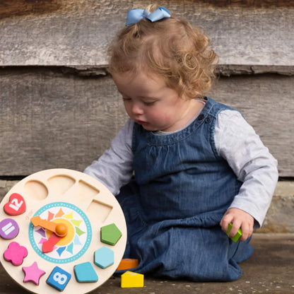 A toddler sits on the ground in a blue denim dress, carefully placing a numbered wooden block into the corresponding slot on a brightly coloured wooden clock.