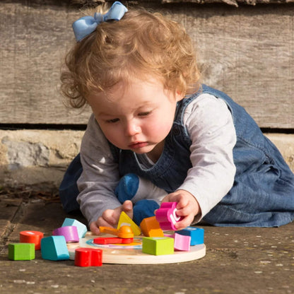 A young child plays on a wooden surface, surrounded by colourful number blocks scattered around a wooden clock toy with rotating hands.