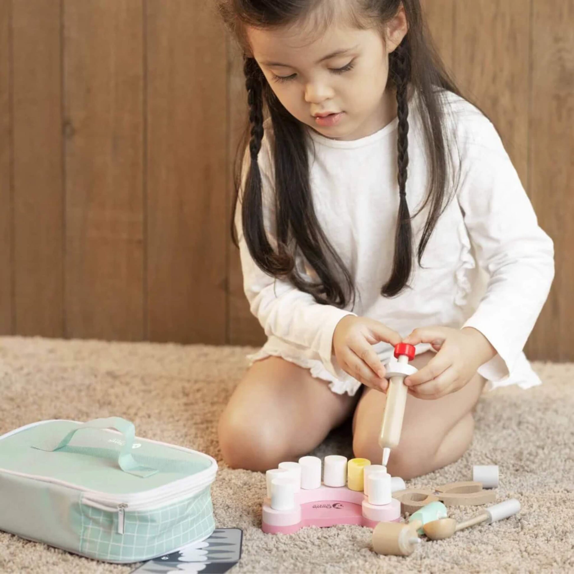 Young child sitting on a carpet, holding a wooden syringe above a pink toy tooth model, with dental tools and a green case beside.