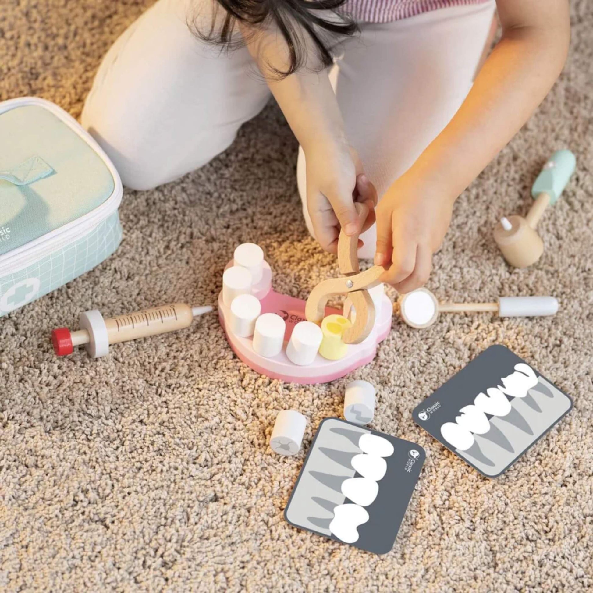 Close-up of a child using toy wooden pliers to grip a yellow play tooth from a pink gum-shaped base, with dental X-ray cards nearby.
