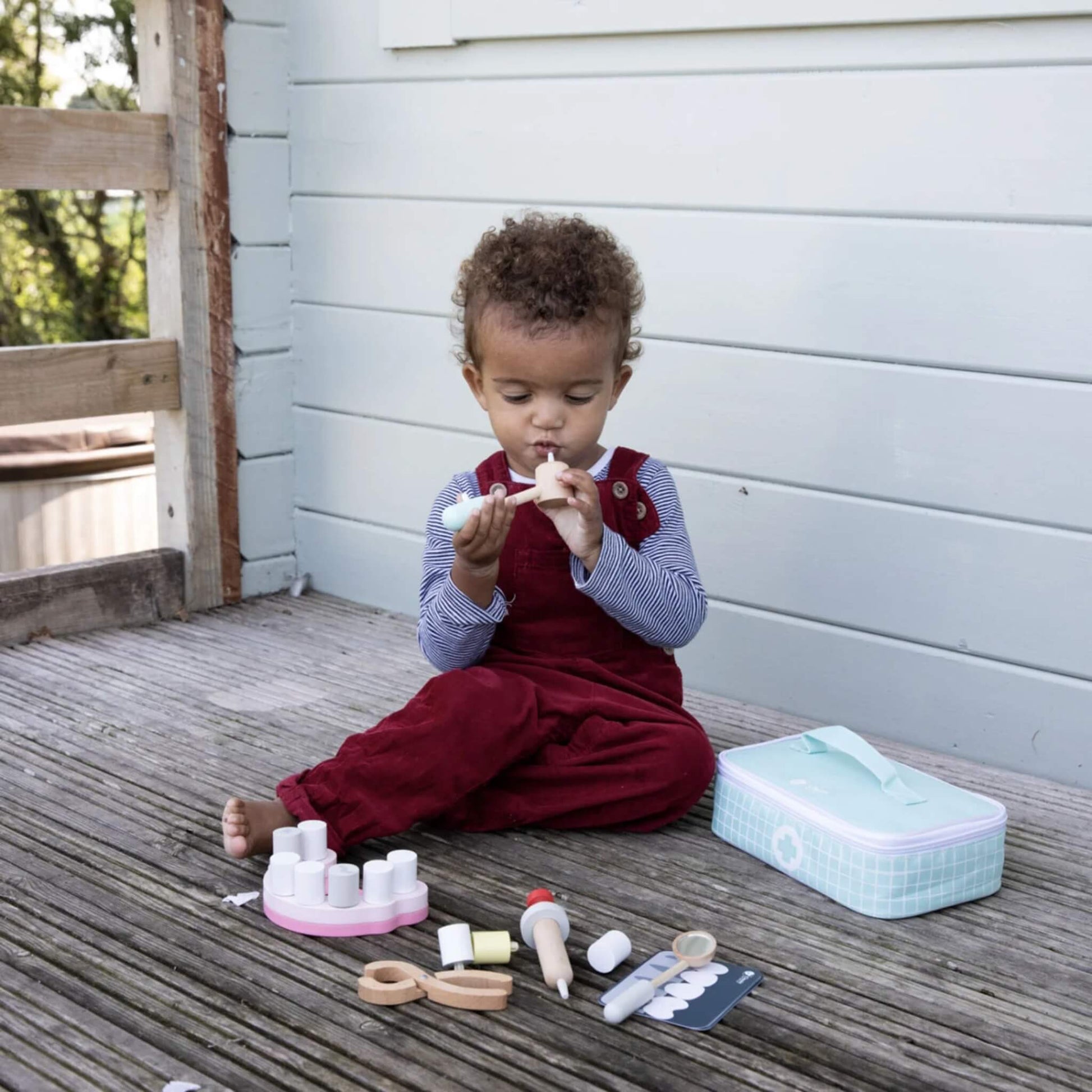 Toddler on a wooden deck, reaching for dental play tools laid out beside a pink tooth model and a pastel green zip-up case.