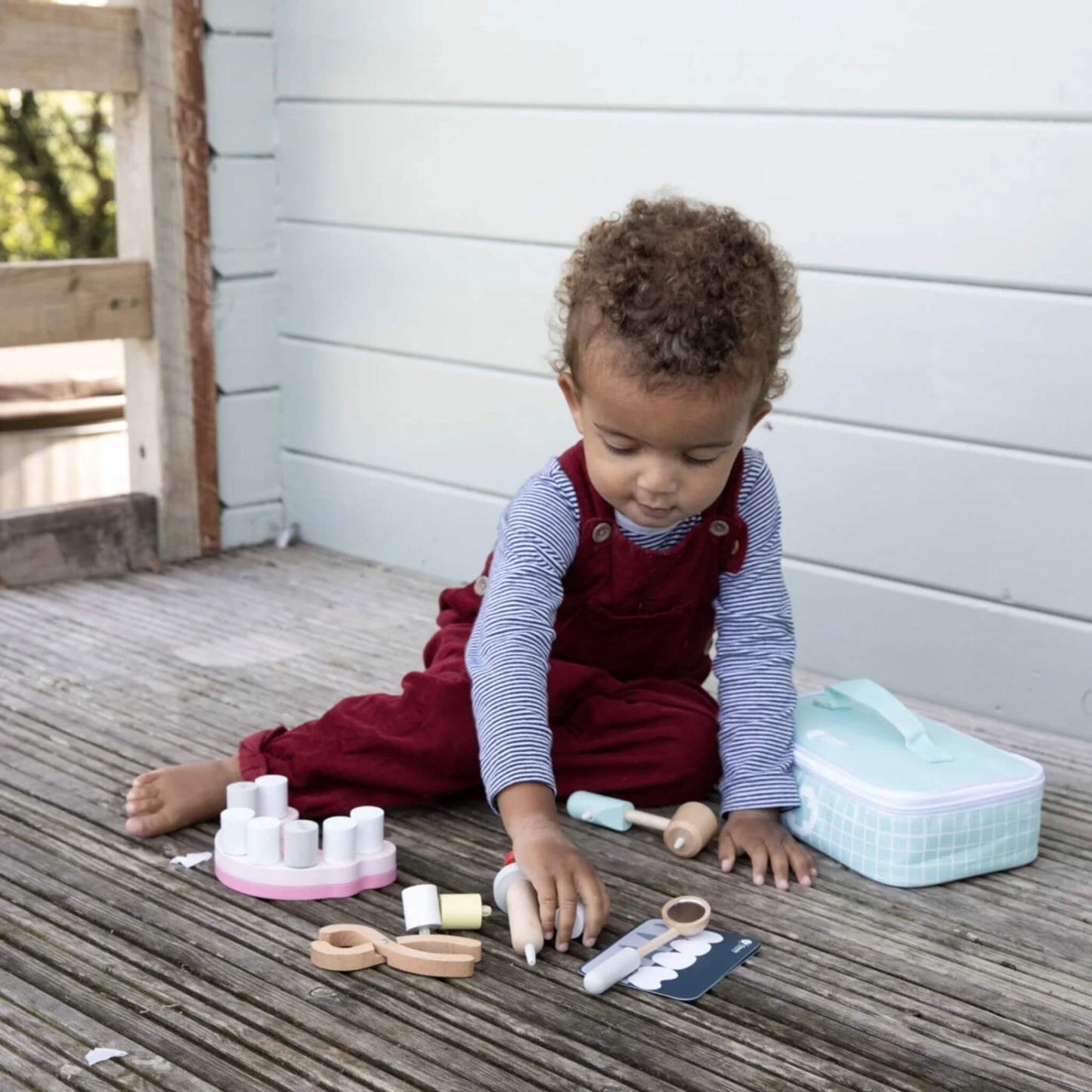 Toddler sitting on wooden decking, wearing a striped top and red dungarees, examining a wooden dental tool with toy teeth and accessories nearby.