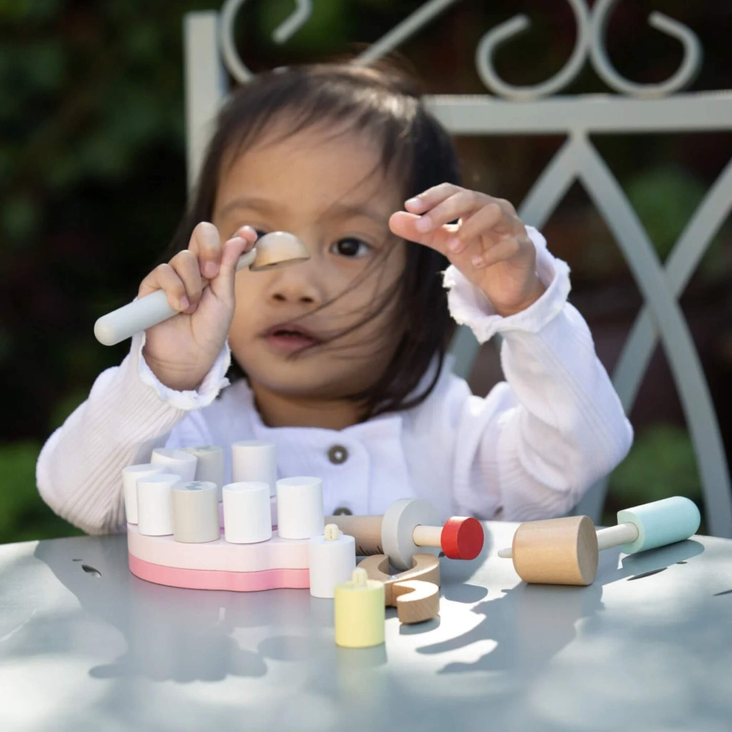 Close-up of a child at a garden table, lifting a wooden dental mirror toward their face, with a pink gum-shaped base and wooden tools in front.