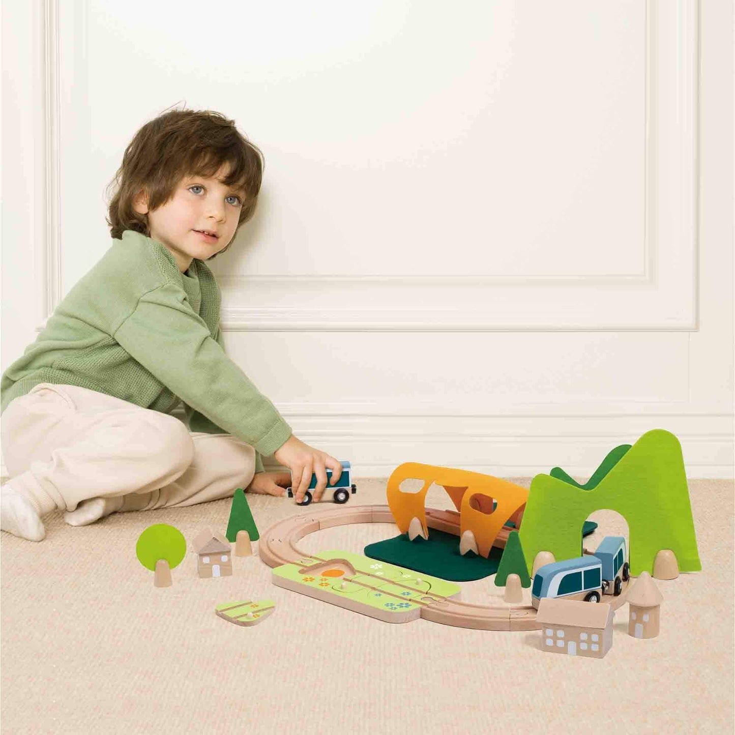 Young child in a green jumper playing with a wooden train set on a beige carpet, pushing the train along the curved track.