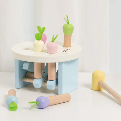 Wooden pounding toy set on a white surface, with radish pieces scattered around and a pastel yellow mallet at the side.