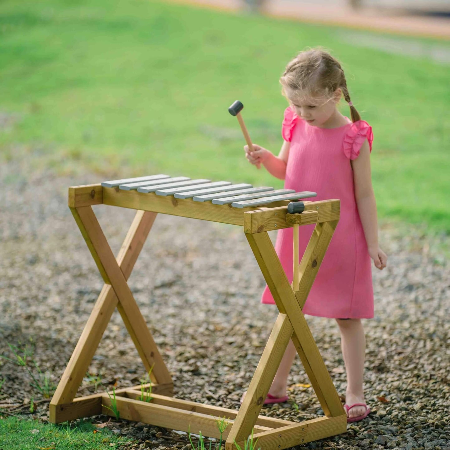 Child in a pink dress playing an outdoor metallophone with a mallet, standing on a gravel surface.