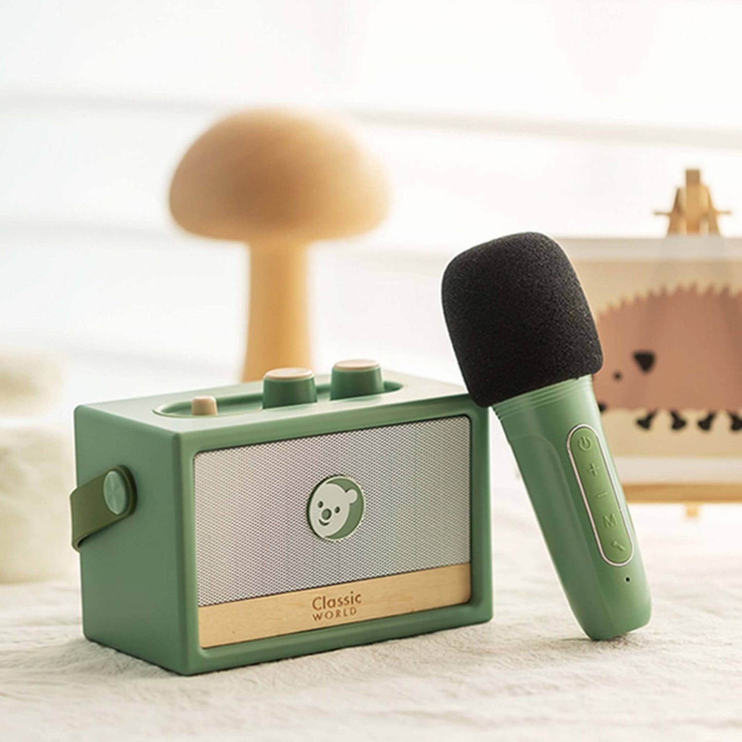 Green toy speaker and handheld microphone set placed on a soft surface with a blurred nursery lamp and wall art in the background.