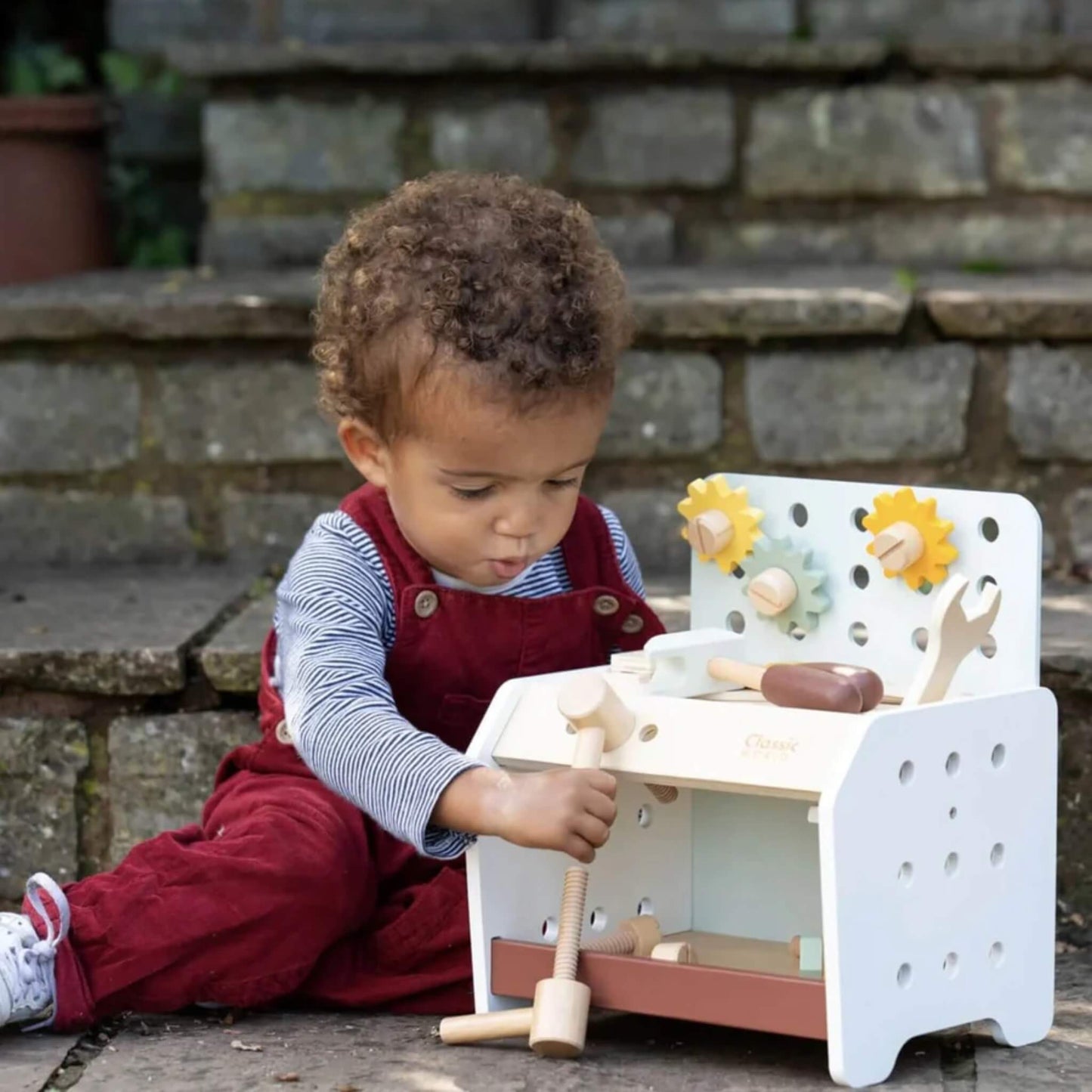 Toddler sitting outdoors on stone steps, playing with a wooden toy mini workbench, focused on twisting wooden bolt into place.