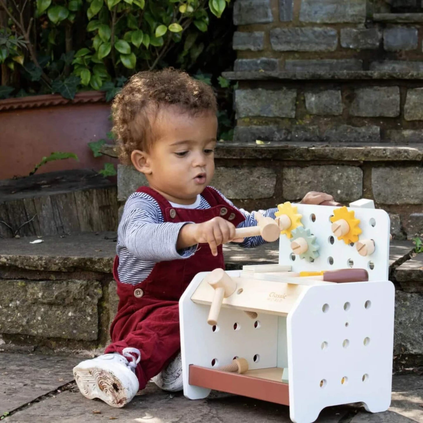 Toddler outdoors using wooden hammer to tap parts on a wooden toy mini workbench, surrounded by toy tools and gears.