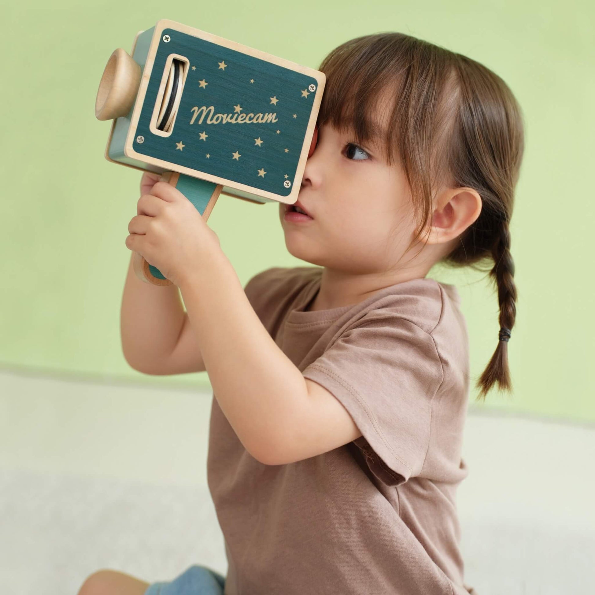 Toddler holding wooden pretend play camera toy and looking through kaleidoscope lens while using handle during imaginative play.
