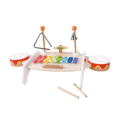 Wooden music table with rainbow xylophone, two small drums, triangle, cymbal, bell, and guiro, shown from side angle with drumsticks resting on floor.
