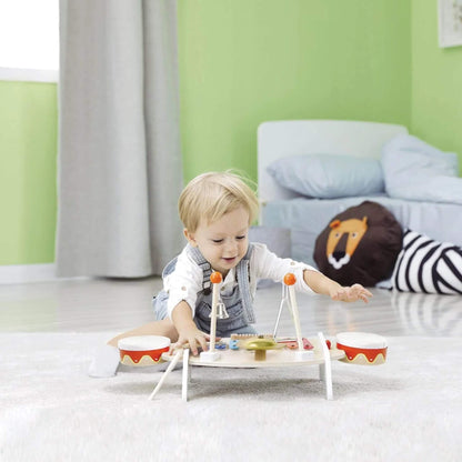 Toddler playing enthusiastically with a wooden music table, sitting on a soft carpeted floor in a bright room with green walls and cosy decor.