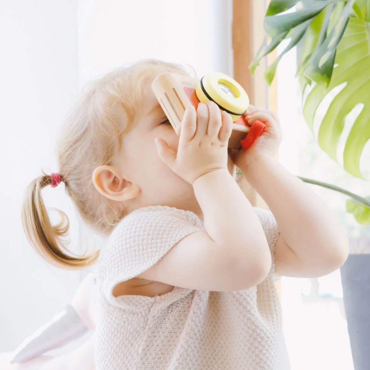 Young child holding wooden toy camera up to face, peeking through viewfinder near a bright window with green houseplant in background.