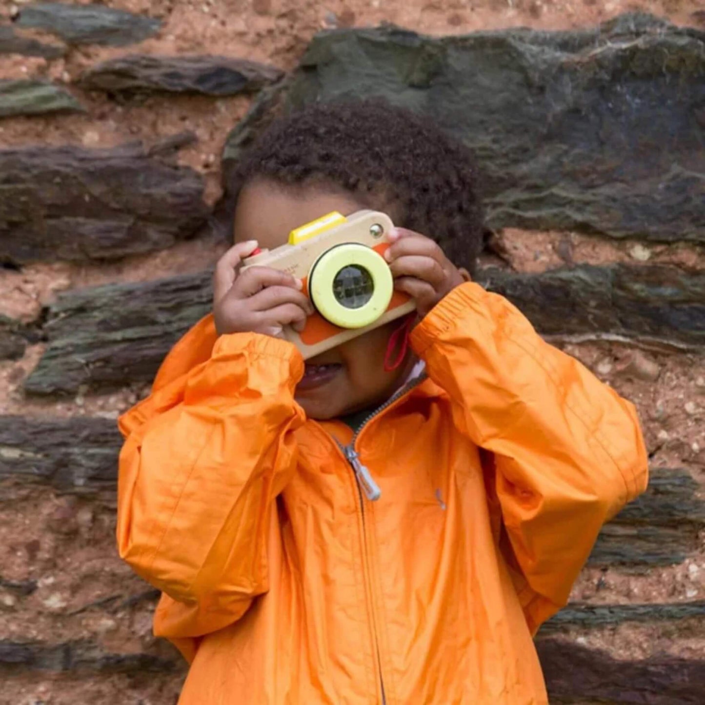Smiling child outdoors in orange raincoat, holding toy camera to face, appearing to “take a picture” against a rustic stone wall backdrop.