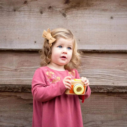 Toddler in pink dress with bow in hair, smiling and holding wooden toy camera with both hands, standing near rustic wooden wall.