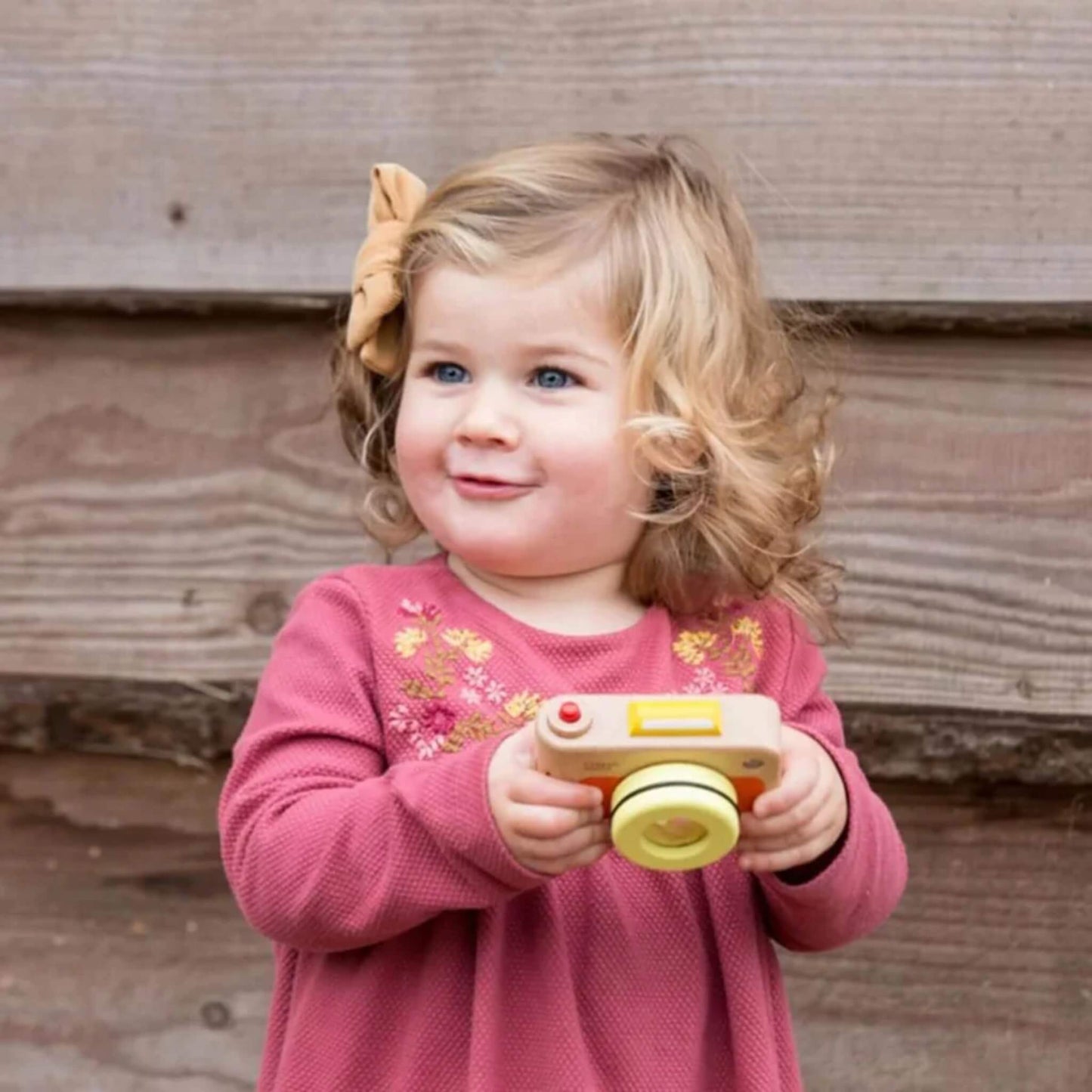 Toddler with wavy blond hair in pink dress holding toy camera, standing against wooden planks, gazing curiously into the distance.